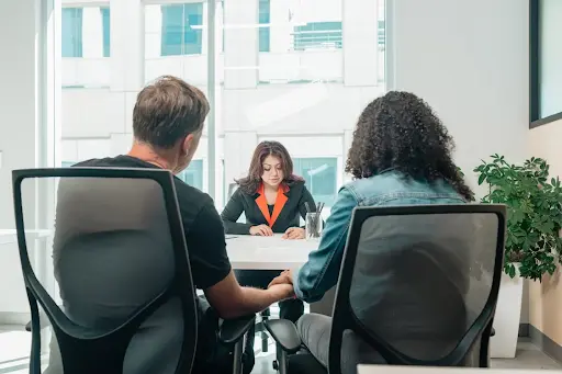 Two people sitting at a desk in an office speaking to one other person on the other side of the desk